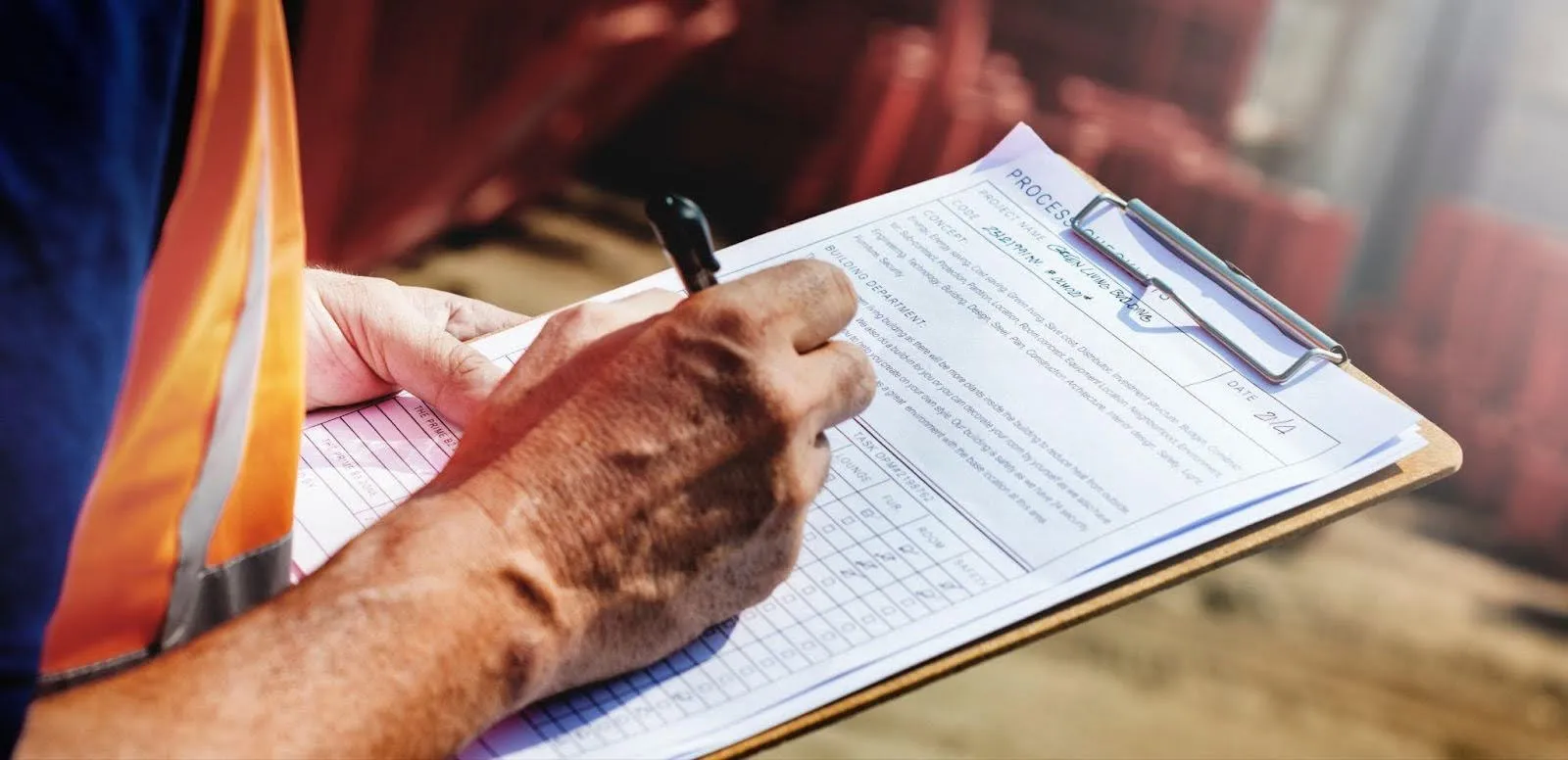 A man wearing an orange vest is focused on writing notes on a clipboard in an outdoor setting