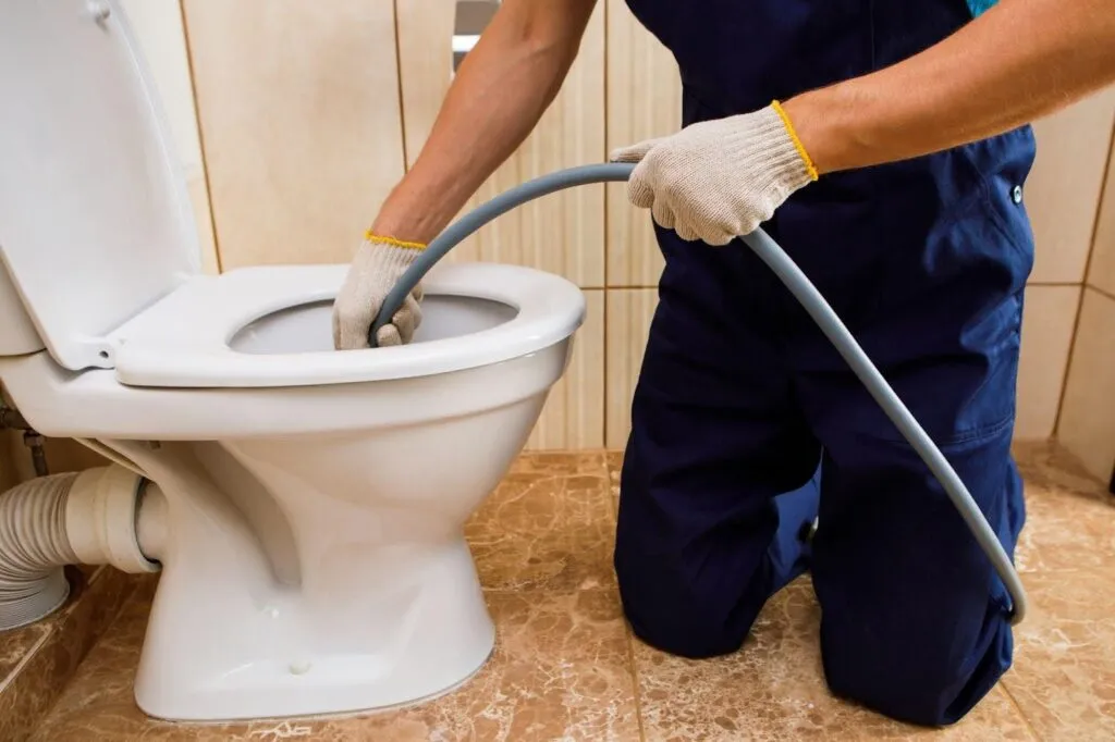 A man in overalls cleans a toilet using a hose demonstrating proper sanitation practices
